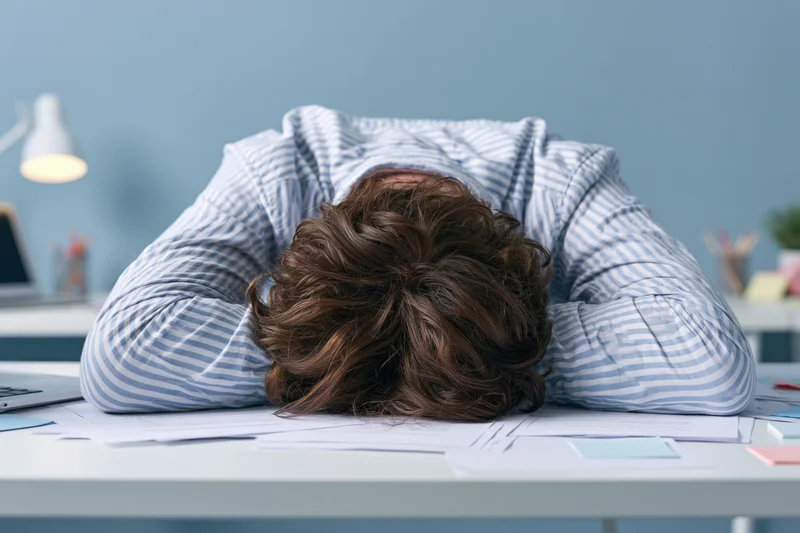 Woman with brown hair slumped face-down on white office desk surrounded by papers and laptop