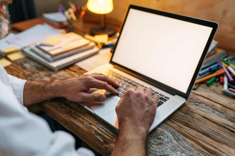Man typing on laptop with blank white screen at wooden desk