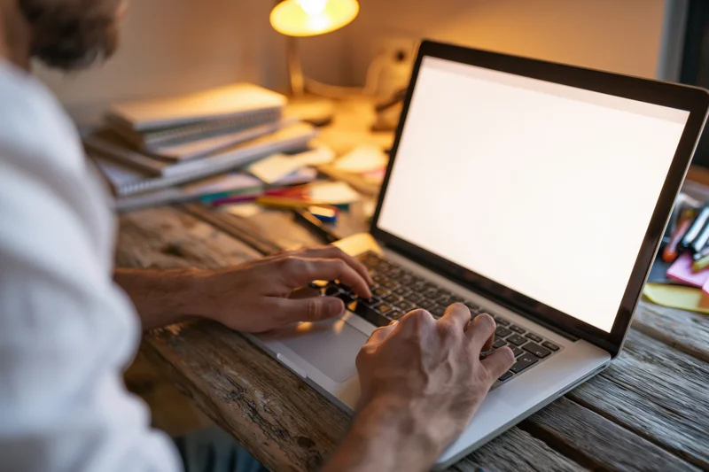 Man typing on laptop with blank white screen at wooden desk