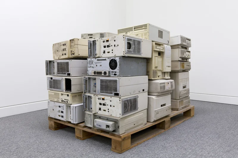 Stack of discarded desktop computer towers on wooden pallet in gallery space