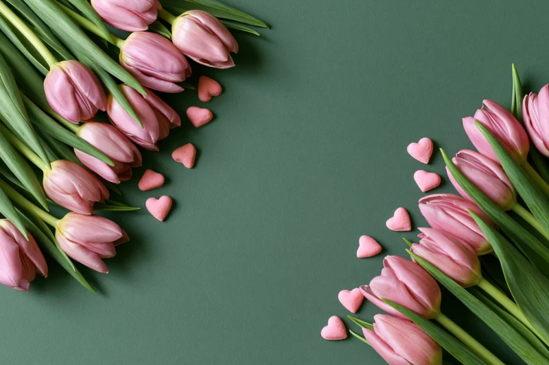 Pink tulips with green stems arranged in two diagonal clusters on dark green surface with scattered heart-shaped candies and central copyspace