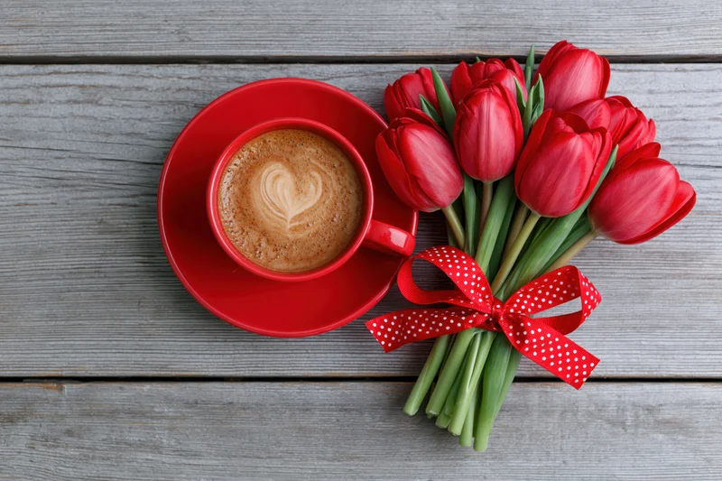 Red ceramic cup of latte with heart art and red tulip bouquet on gray wooden table