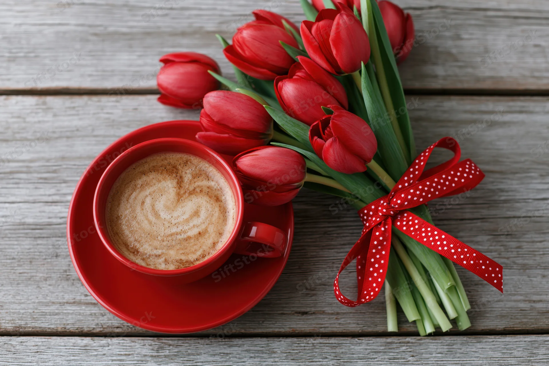 Red ceramic cup of latte art coffee with heart design beside a bouquet of red tulips tied with polka dot ribbon on rustic wooden table
