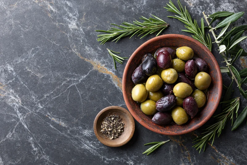 Mixed green and dark olives in a rustic clay bowl with rosemary and pepper on a gray marble surface