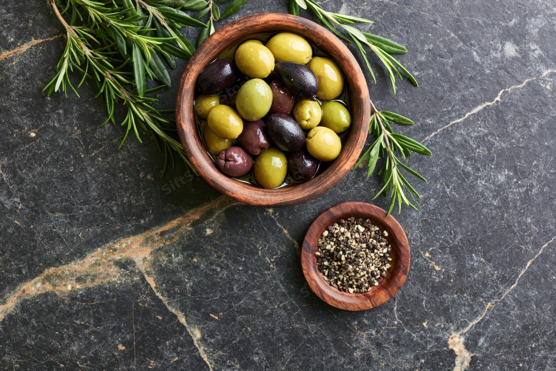 Mixed green and black olives in a wooden bowl with rosemary and ground pepper on a dark stone surface