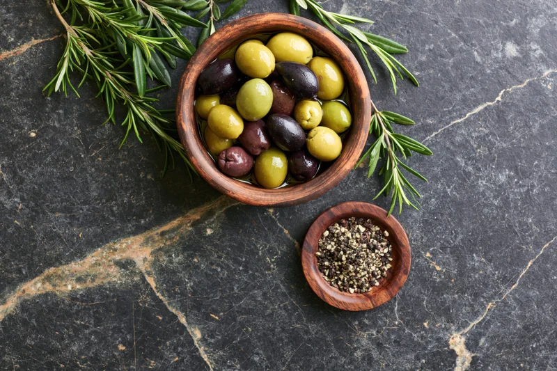 Mixed green and black olives in a wooden bowl with rosemary and ground pepper on a dark stone surface