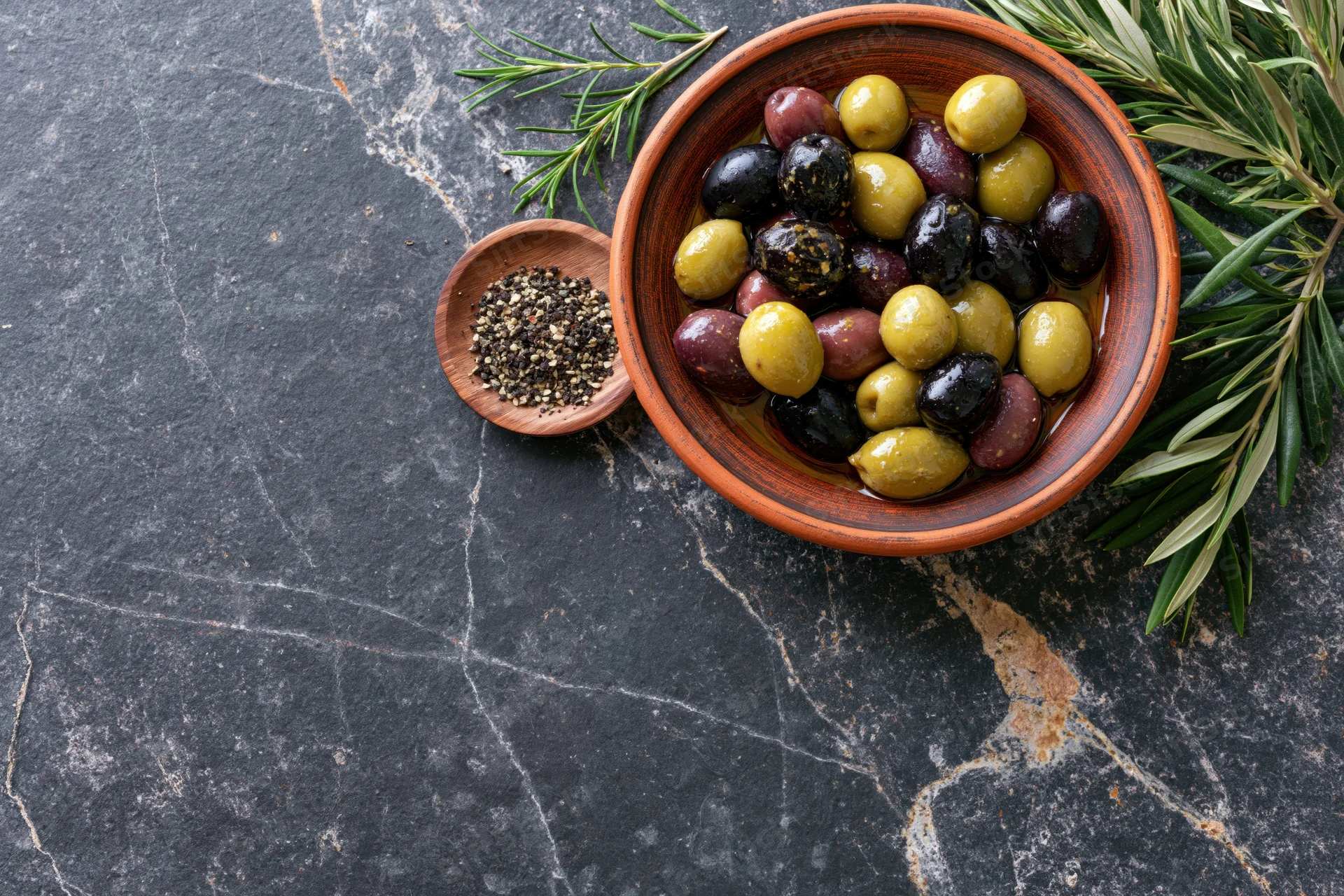 Mixed olives in a clay bowl with rosemary and pepper on a dark stone surface