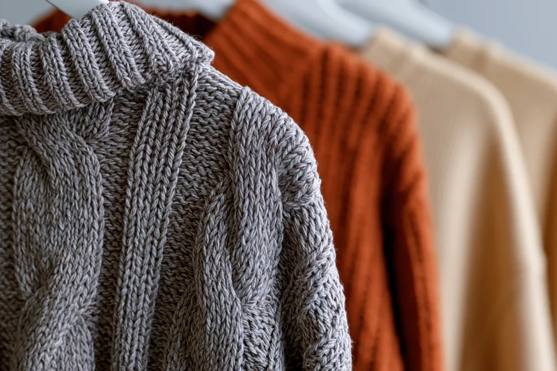 Closeup of gray cable-knit sweater hanging on rack with orange and beige sweaters behind