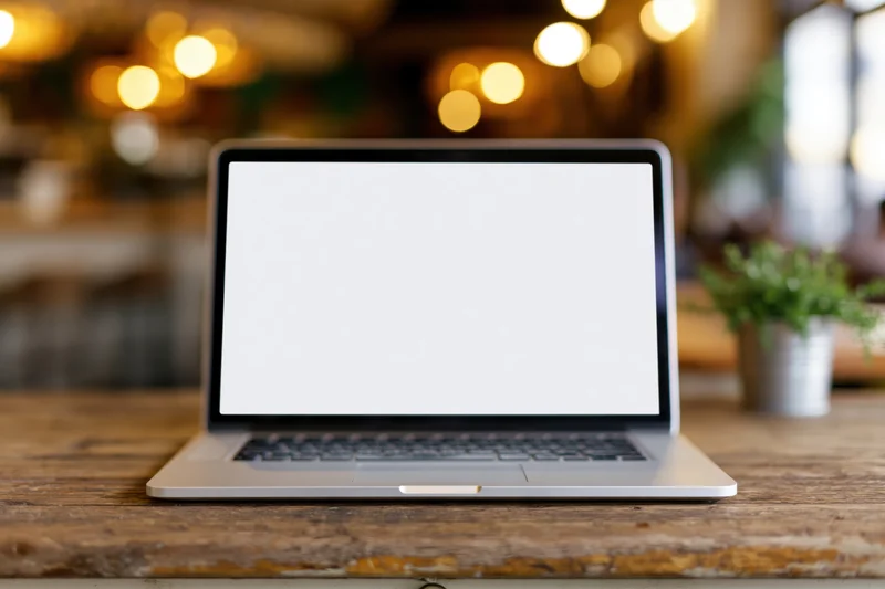 Silver laptop with blank white screen open on wooden desk in cozy interior with warm bokeh lights and small potted plant nearby