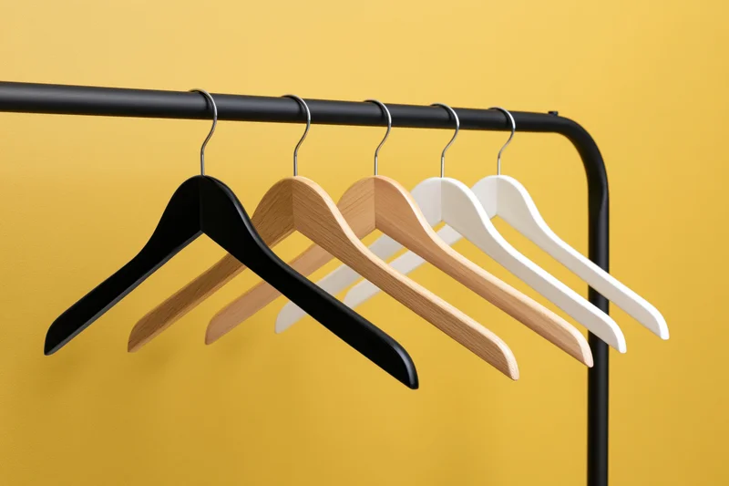 Five empty clothes hangers in black, natural wood, and white hanging on a black metal rack against a yellow background
