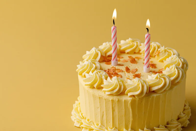 Small pale yellow birthday cake with two lit striped candles on a warm yellow background