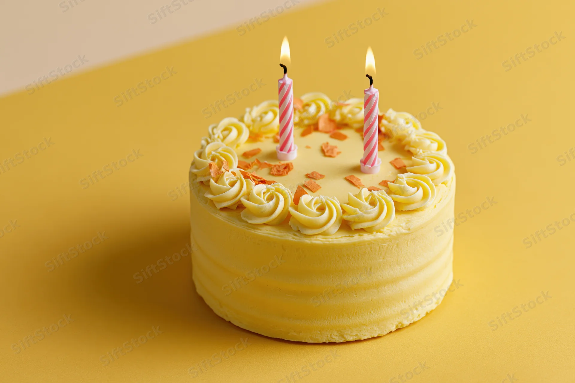 Small pale yellow birthday cake with two lit striped candles on a yellow surface