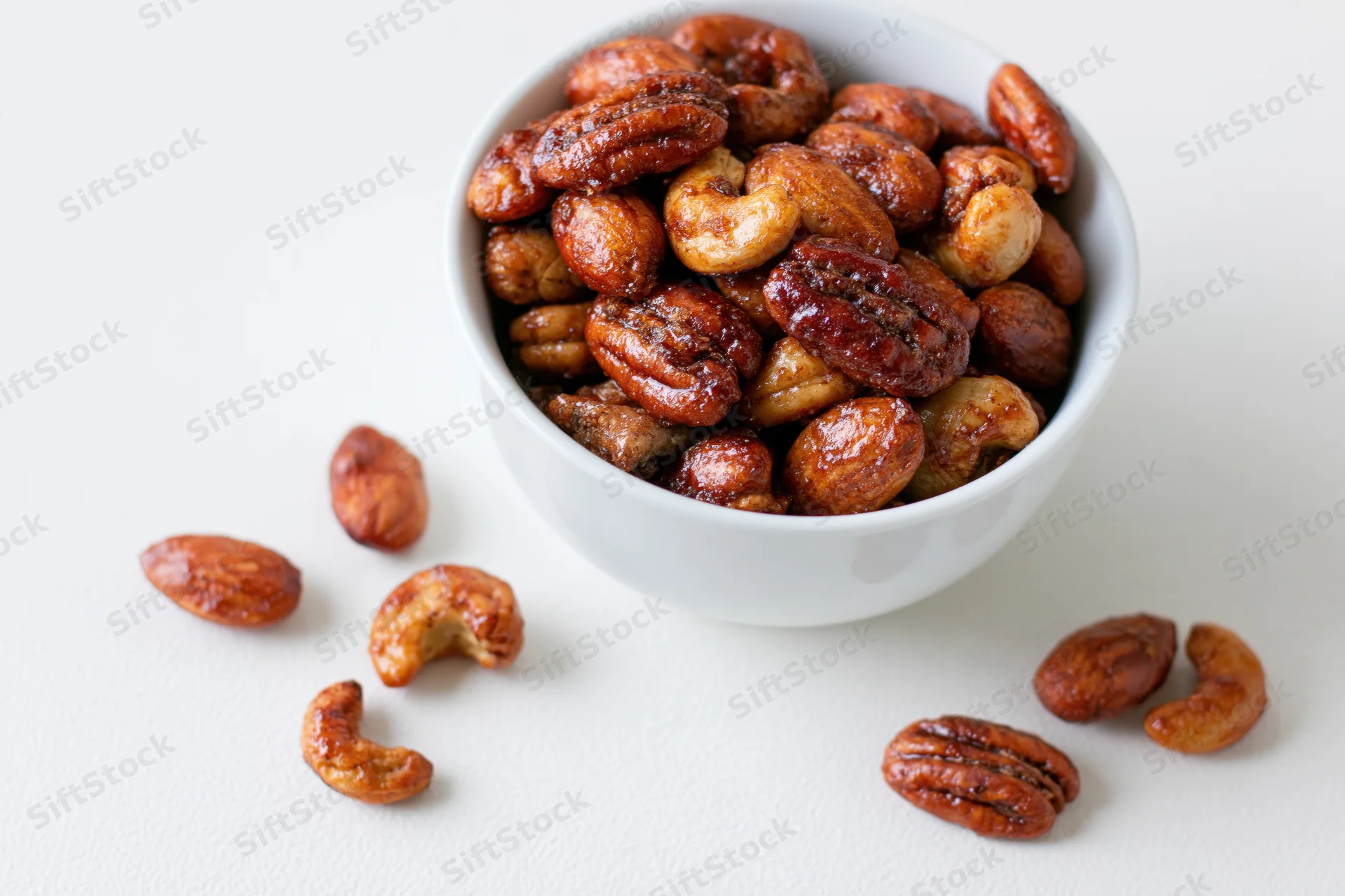 Candied spiced mixed nuts in a white ceramic bowl with scattered pieces on a plain surface