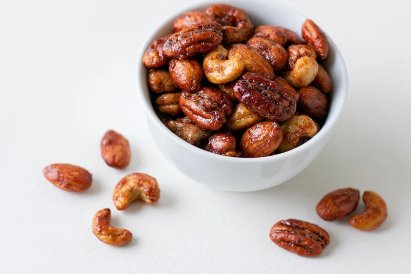 Candied spiced mixed nuts in a white ceramic bowl with scattered pieces on a plain surface