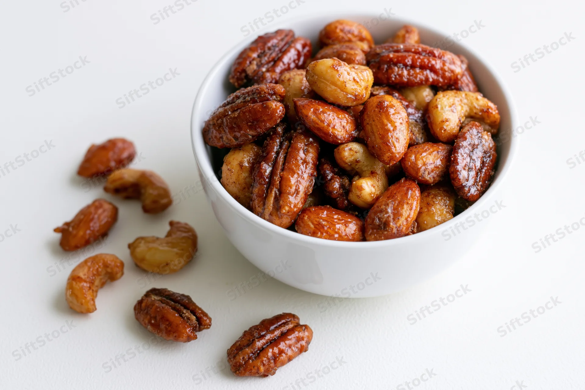 Candied spiced mixed nuts in a white ceramic bowl with scattered pieces on a plain surface
