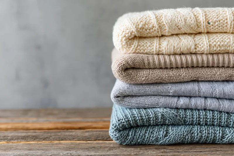 Stack of four folded knit sweaters in neutral tones on a rustic wooden table