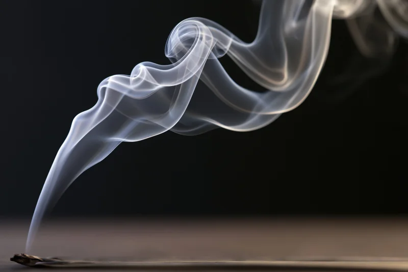 Thin white smoke curling upward from an incense stick on a dark surface