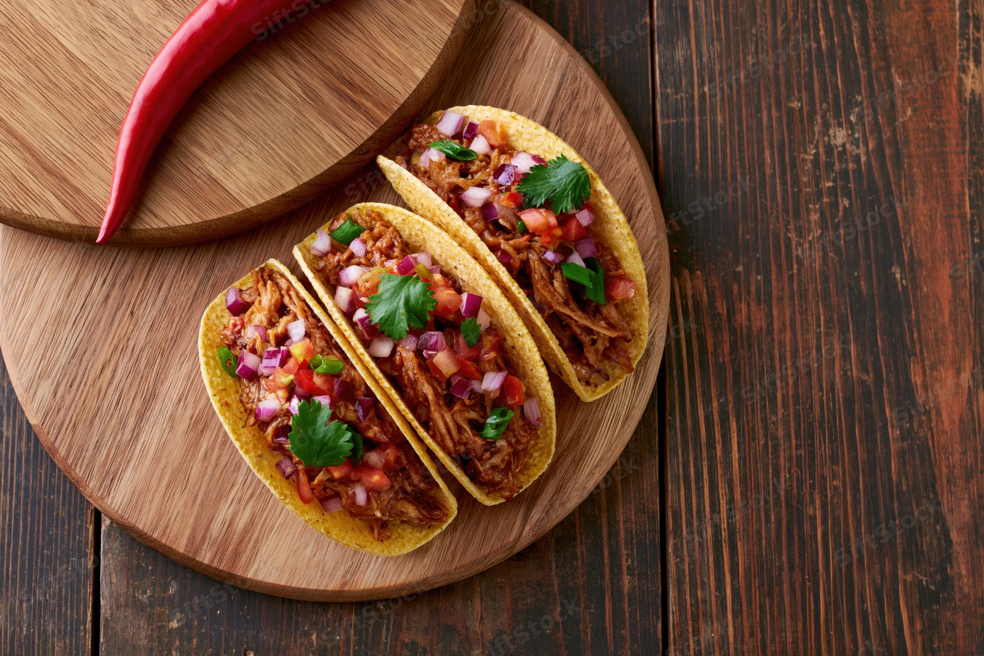 Three hard shell tacos filled with pulled meat and diced vegetables on a wooden board