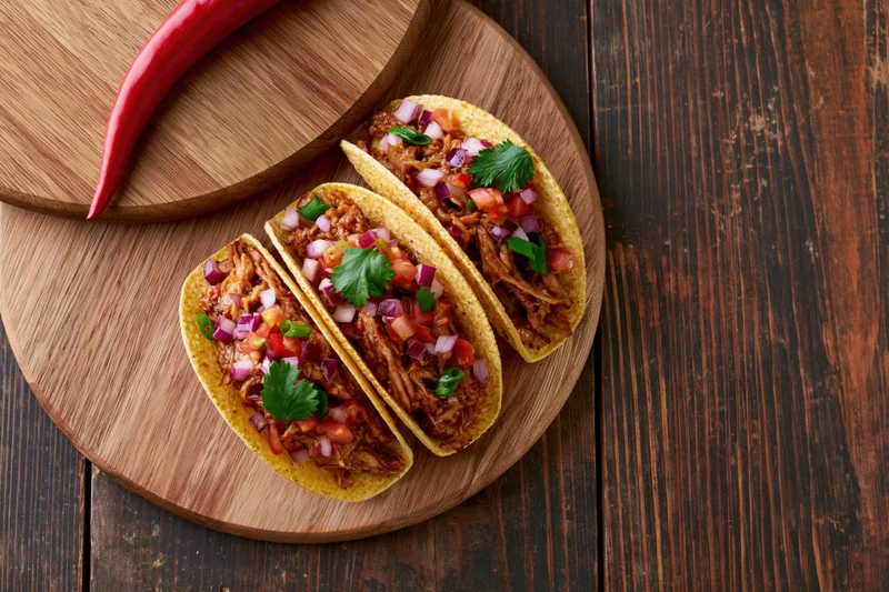 Three hard shell tacos filled with pulled meat and diced vegetables on a wooden board