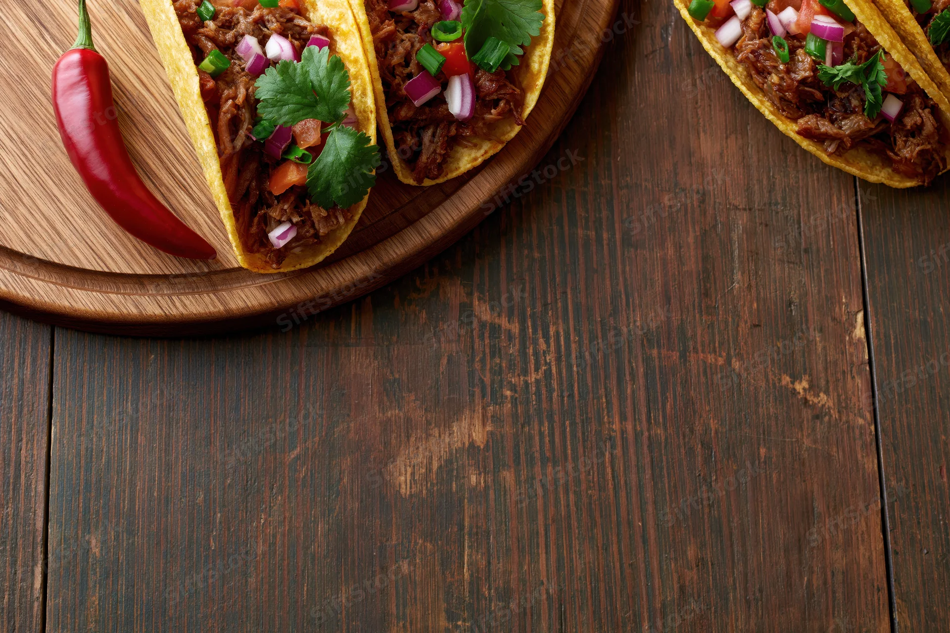 Three hard shell tacos filled with pulled shredded meat and diced vegetables on a wooden board over a dark rustic table