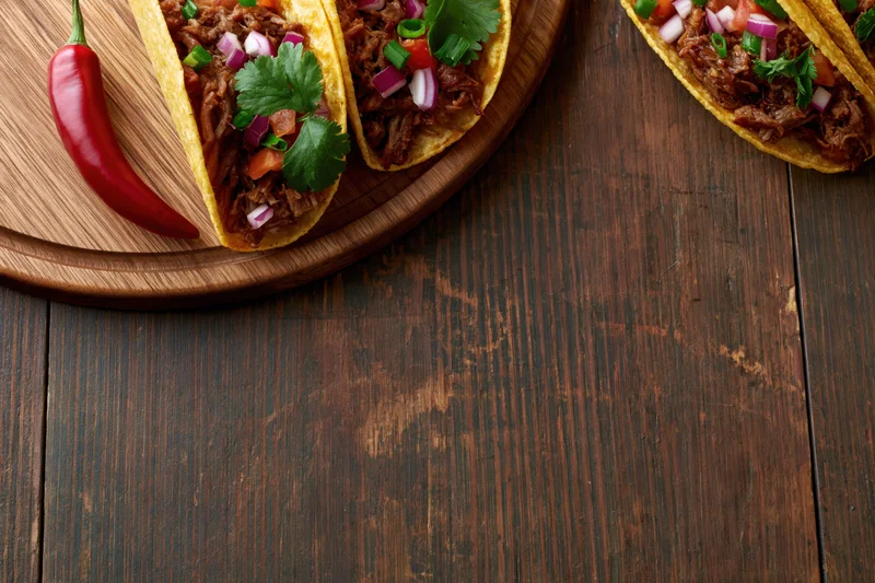 Three hard shell tacos filled with pulled shredded meat and diced vegetables on a wooden board over a dark rustic table