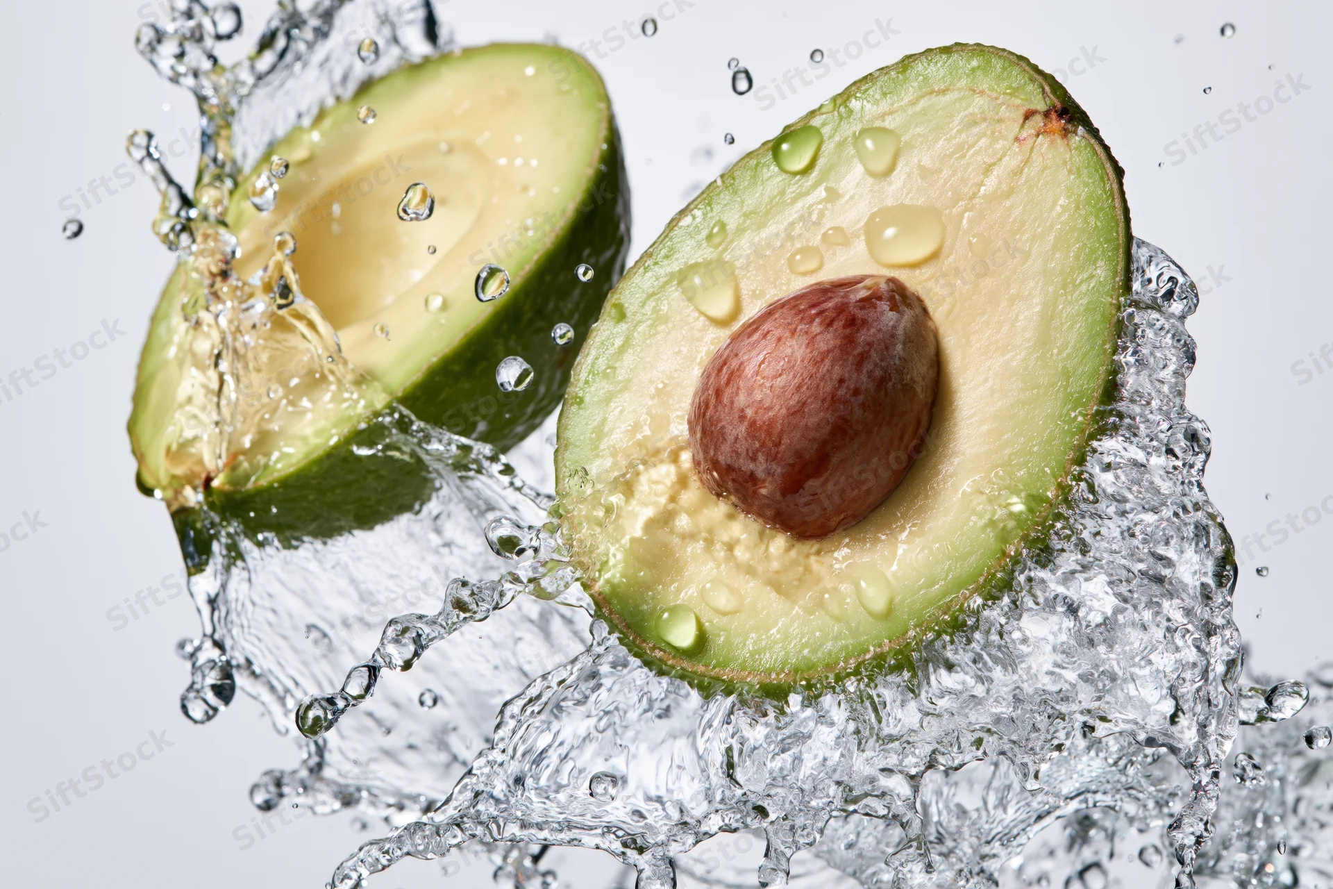 Two avocado halves midair surrounded by dynamic water splash against white background