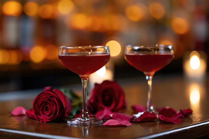 Two coupe glasses with red cocktails on a wooden bar surrounded by red roses and petals under warm candlelight