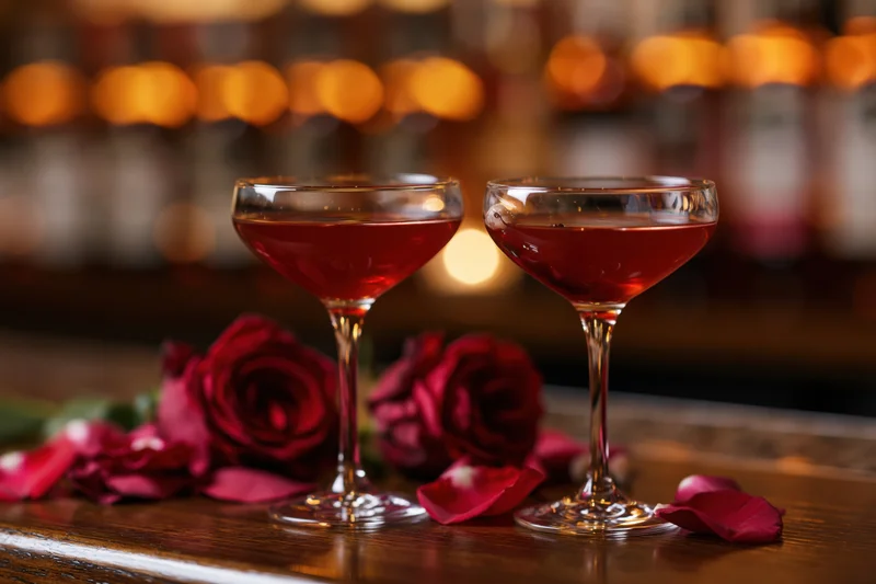 Two coupe glasses with red cocktails on a wooden bar table surrounded by red roses and petals