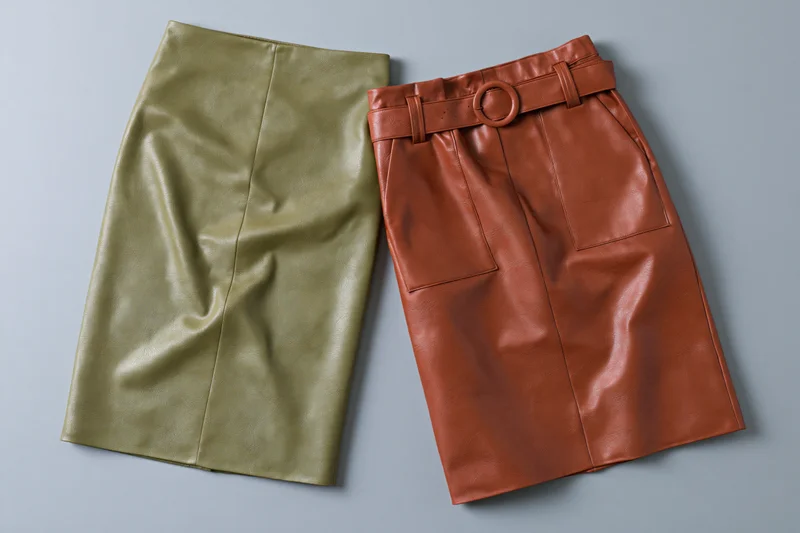 Two faux leather skirts laid flat on a grey surface, one olive and one brown, with belt and pockets