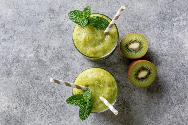 Two glasses of green kiwi smoothie with paper striped straws and mint garnish on gray countertop