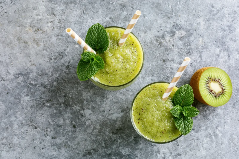 Two glasses of green kiwi smoothie with paper striped straws and mint garnish on a gray stone surface