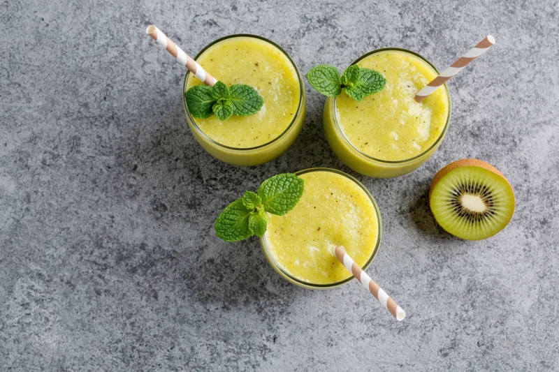 Three glasses of yellow kiwi smoothie with mint garnish and paper straws on gray countertop