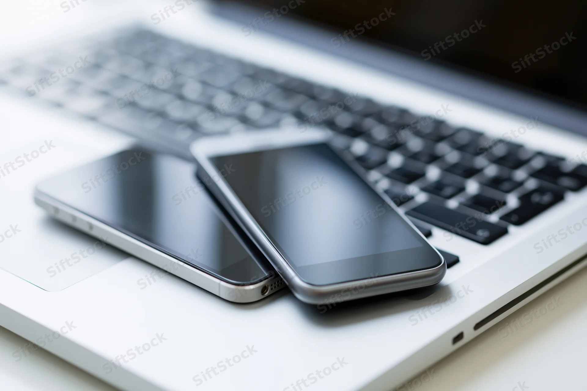 Two smartphones with black screens resting on a silver laptop keyboard