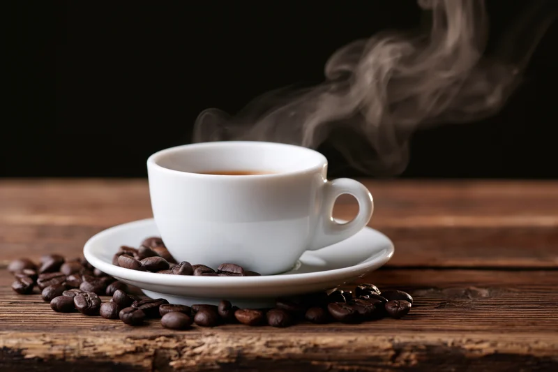 Steaming white ceramic espresso cup on saucer with coffee beans scattered on wooden table