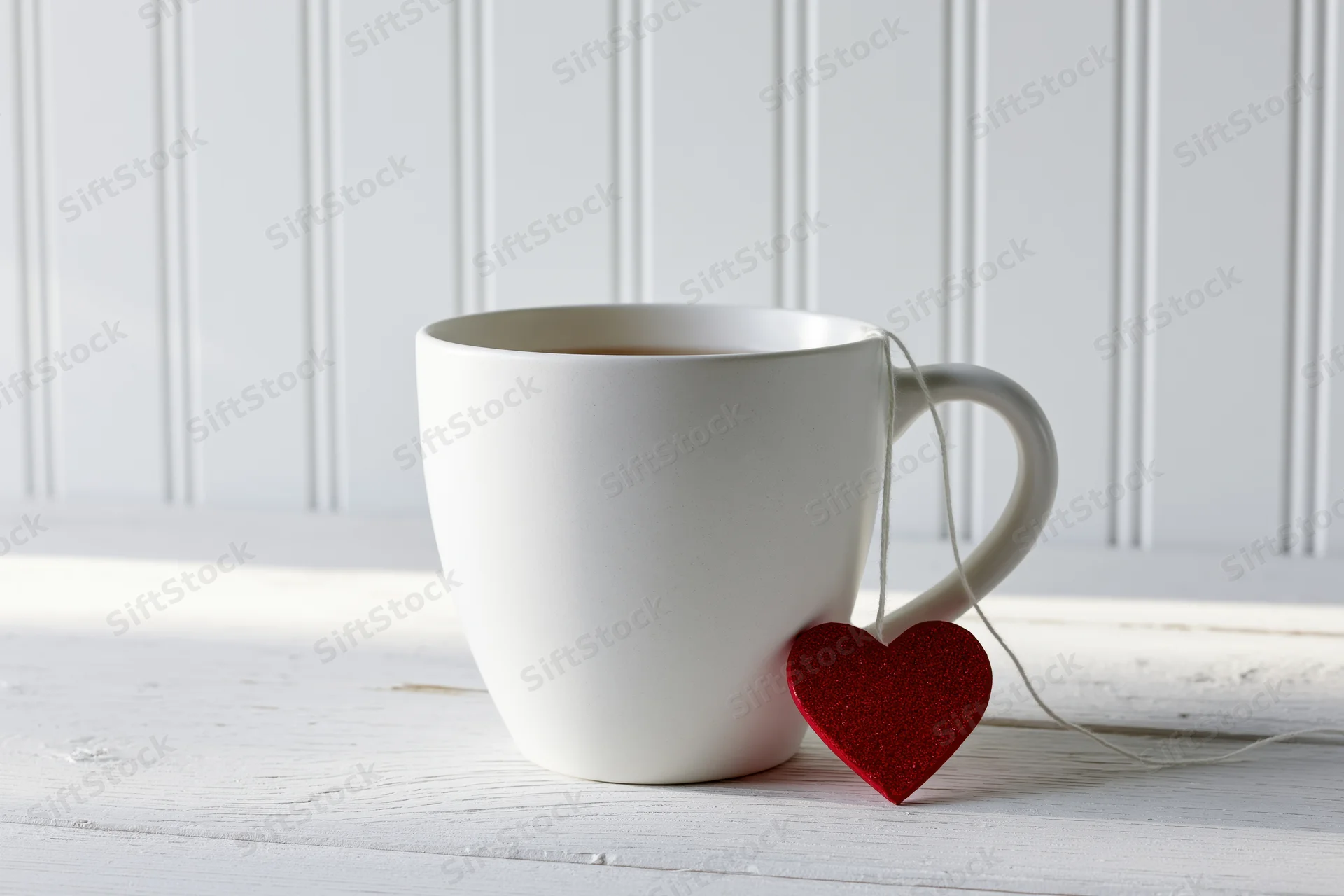 White ceramic mug with tea bag string and red glitter heart tag on wooden surface near window