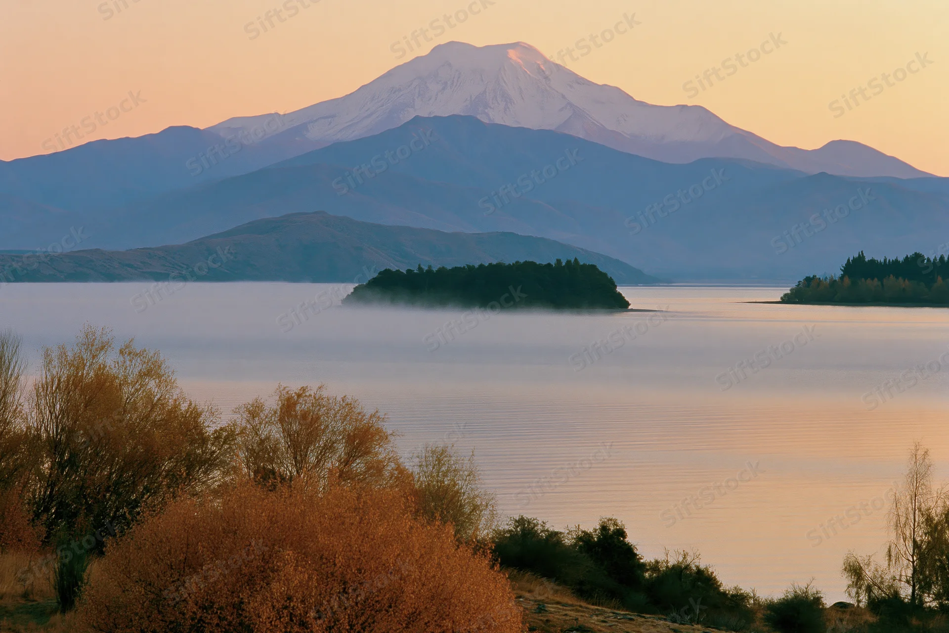 Calm lake at dawn with snow-capped mountain in background, mist hovering over water, autumn trees in foreground
