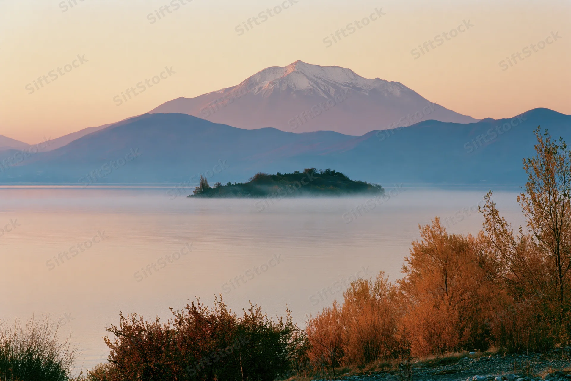 Calm lake at dawn with snow-capped mountain in background, mist rising over water, autumn trees in foreground