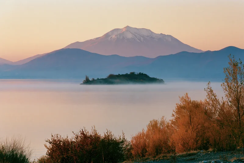 Calm lake at dawn with snow-capped mountain in background, mist rising over water, autumn trees in foreground