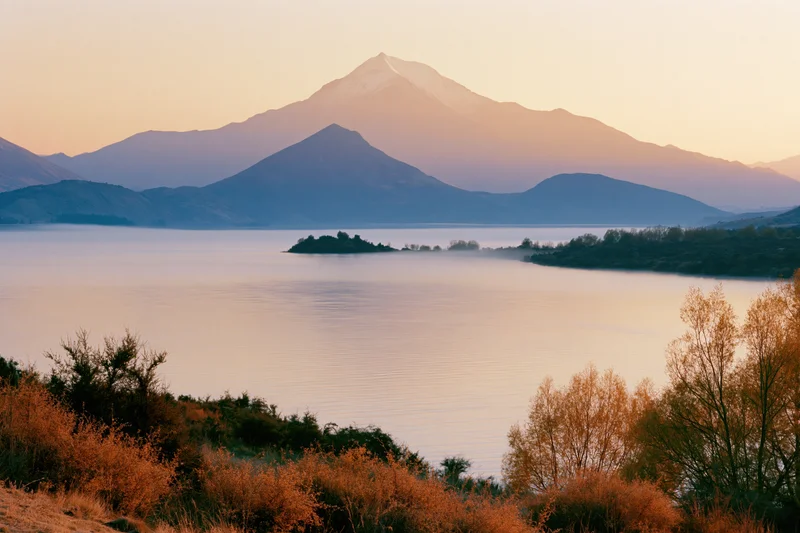 Calm lake reflecting snow-capped mountain at dawn with autumn trees in foreground