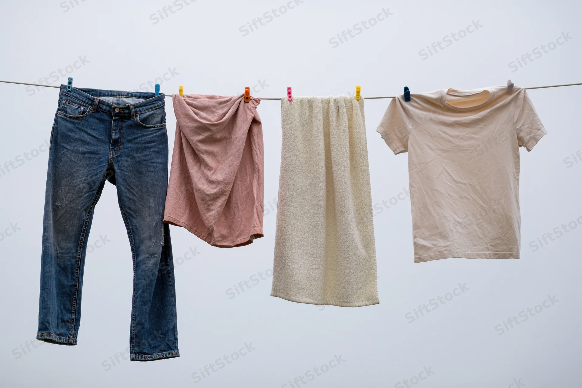 Blue jeans, pink cloth, cream towel and beige tshirt hanging on a clothesline against a light sky