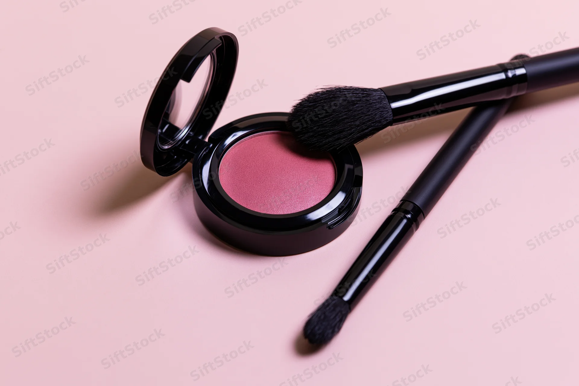 Open blush compact with two black makeup brushes on a pink surface
