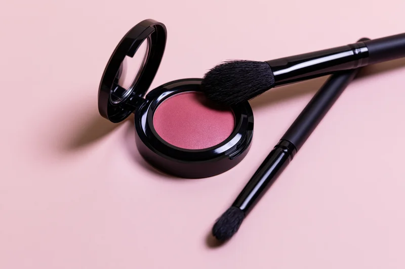 Open blush compact with two black makeup brushes on a pink surface