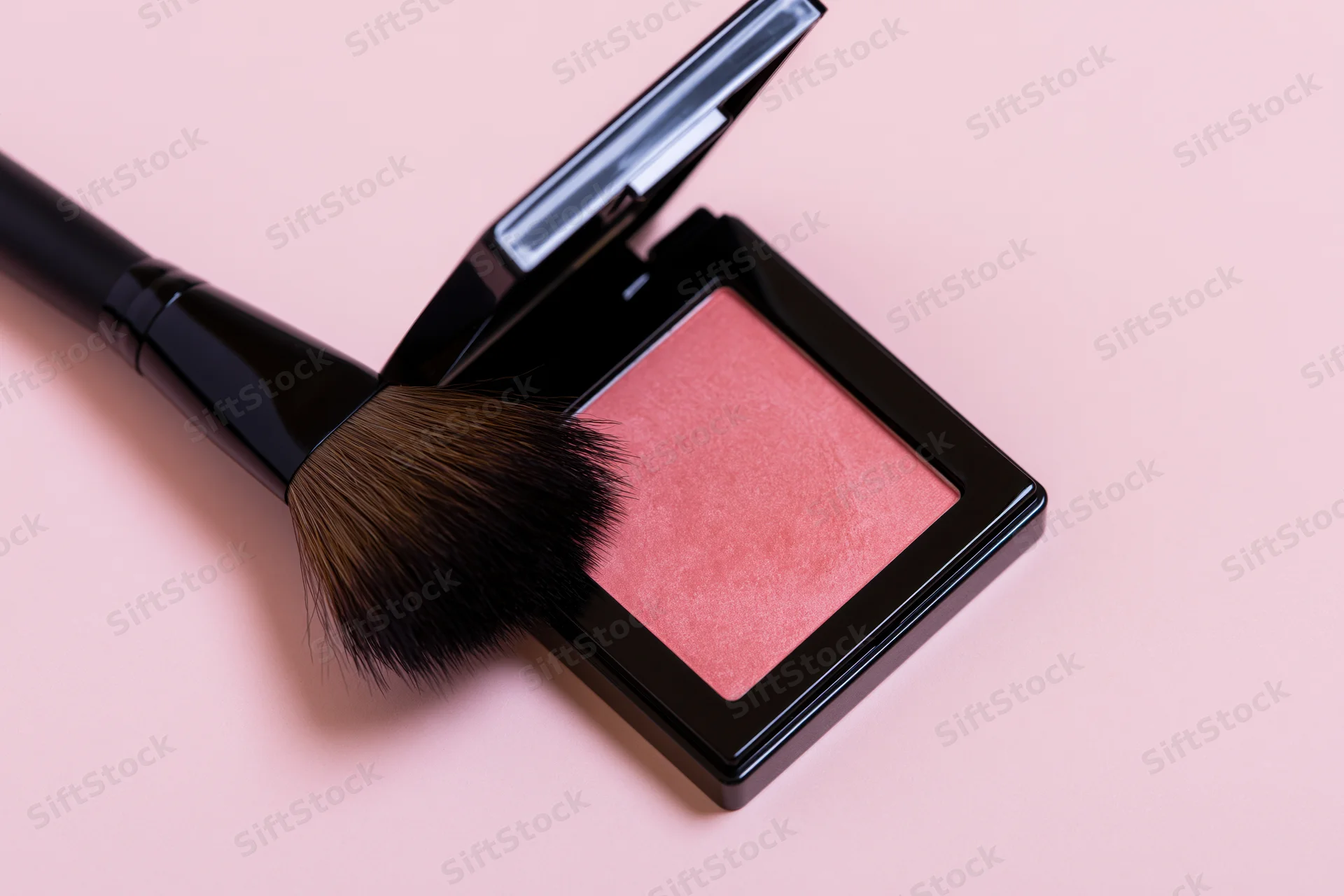 Open blush compact with makeup brush resting on it on a pink surface