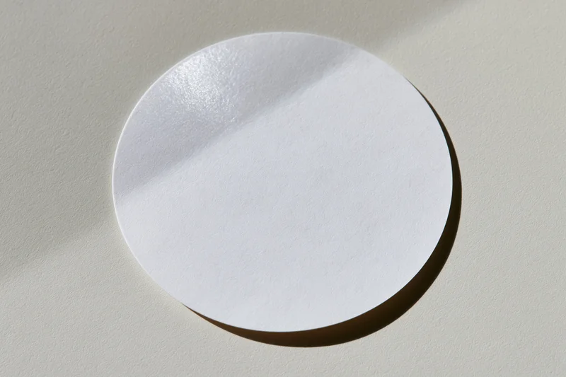 Blank white circular sticker lying flat on a light warm beige textured surface