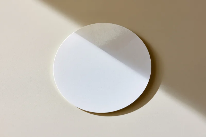 Blank white circular sticker lying flat on a beige surface with soft shadow and copyspace