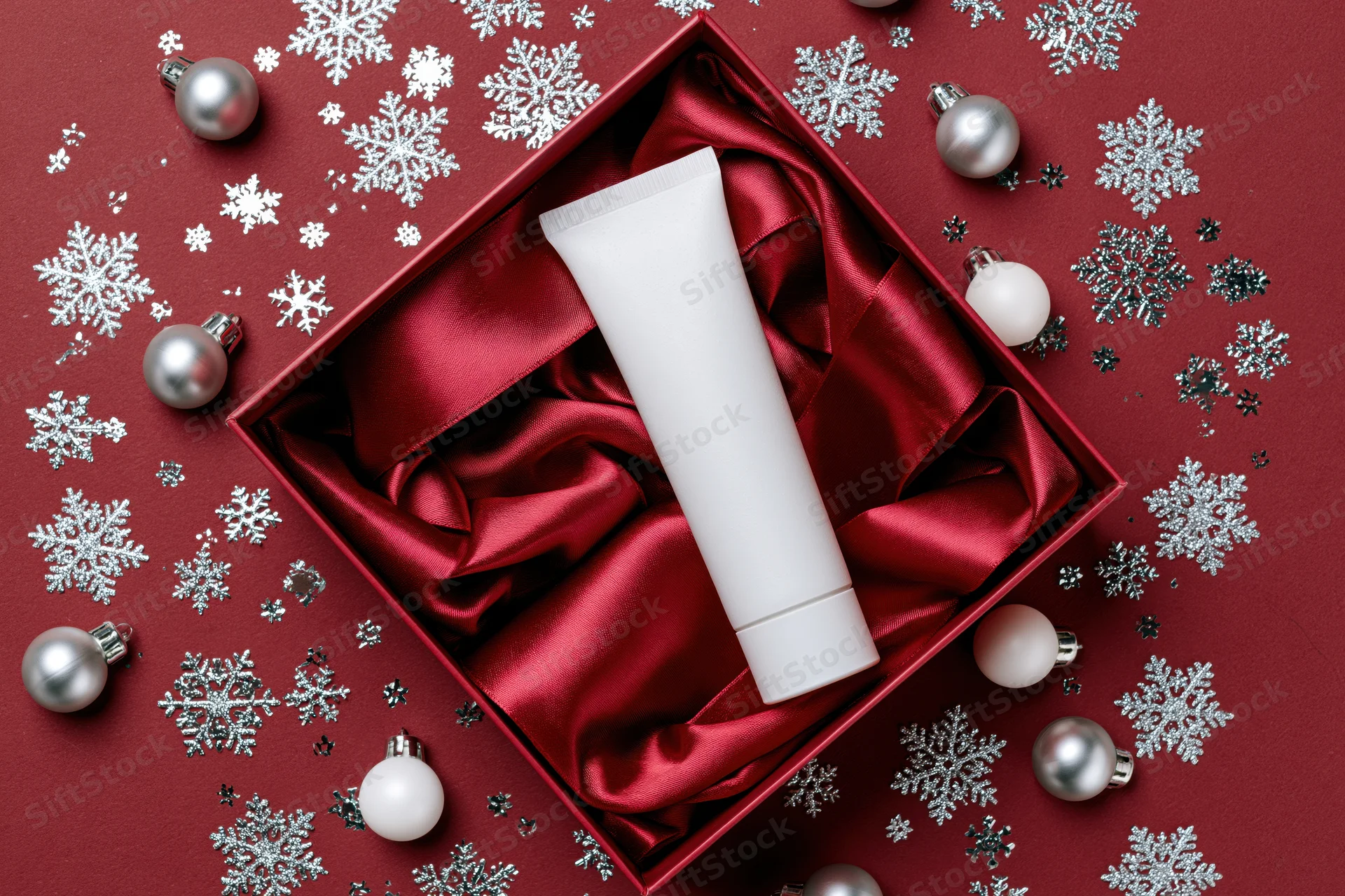 Blank white skincare tube in red satin-lined gift box surrounded by silver snowflakes and ornaments on red background
