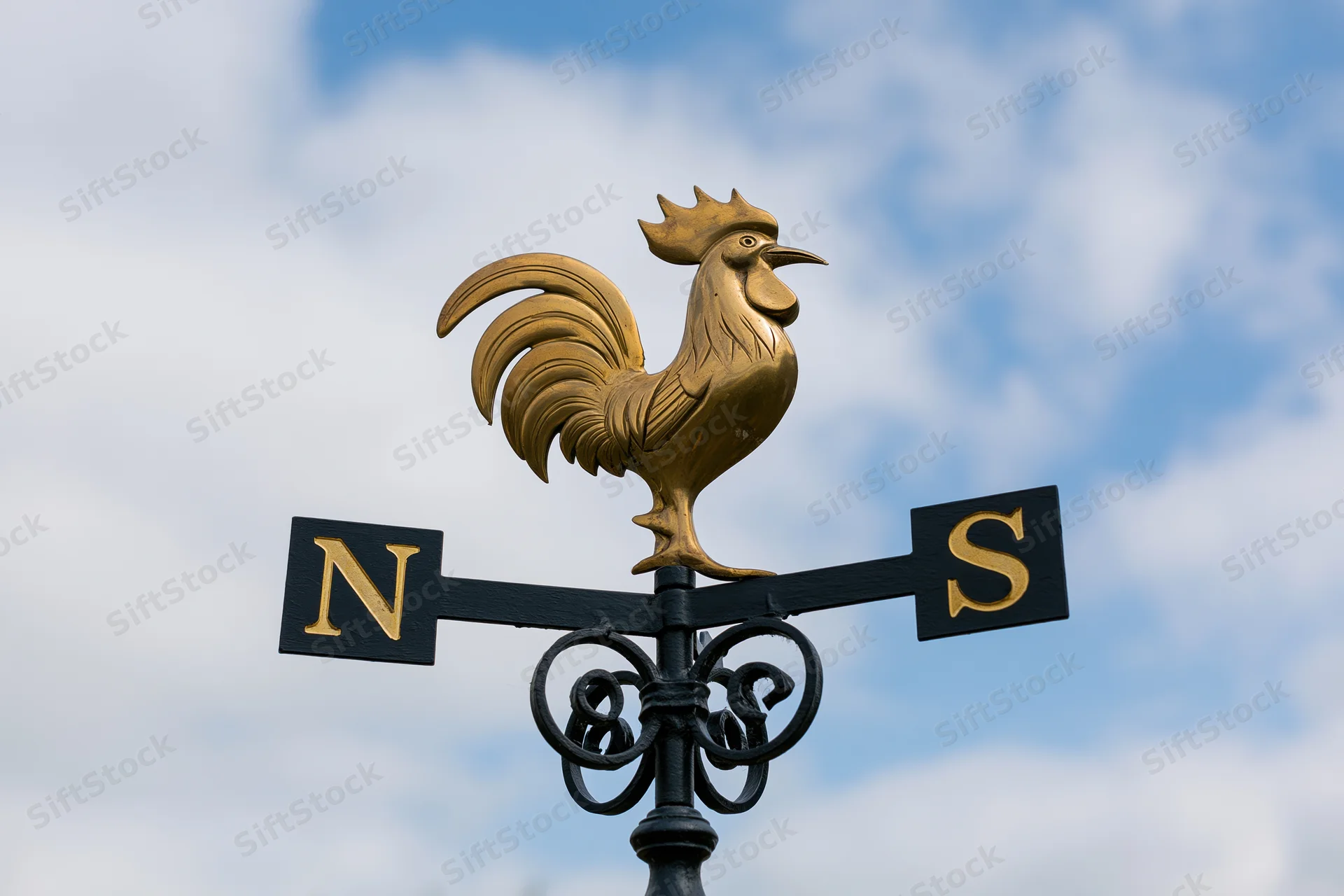 Brass rooster weather vane mounted on wrought iron scroll against blue sky with clouds