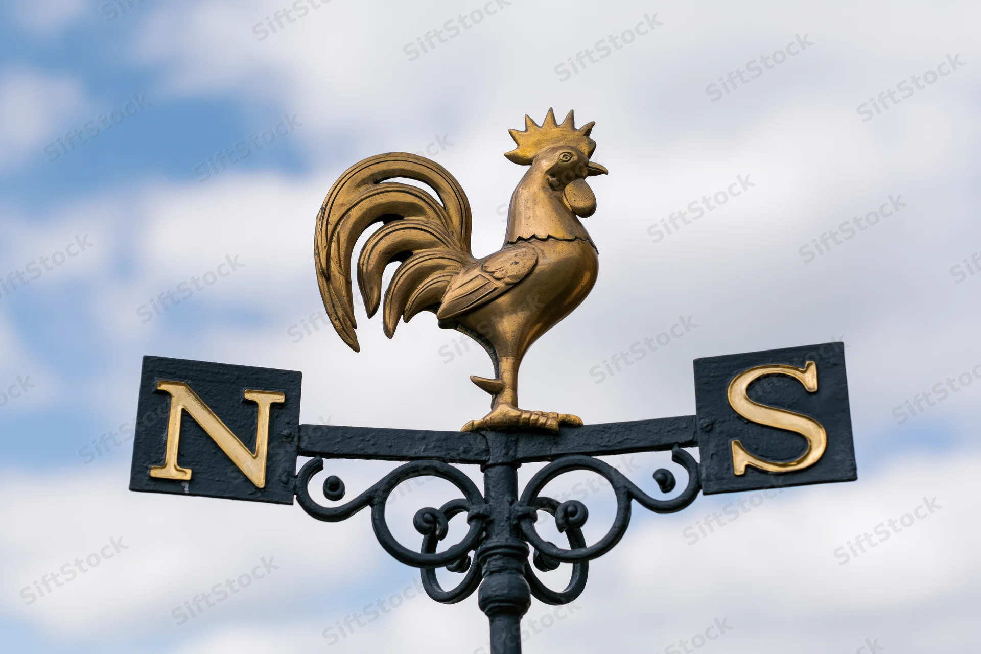 Brass rooster weather vane mounted on wrought iron scroll against cloudy sky