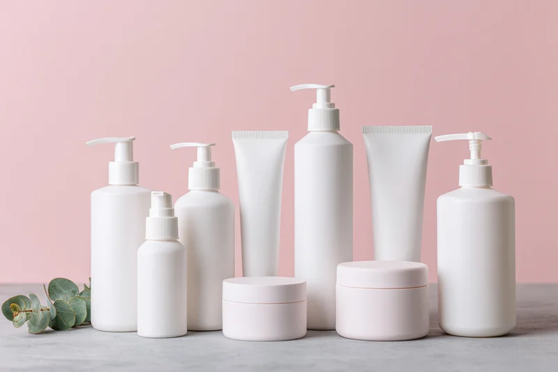 White blank skincare bottles and tubes arranged on a concrete surface with eucalyptus leaves against a pink background