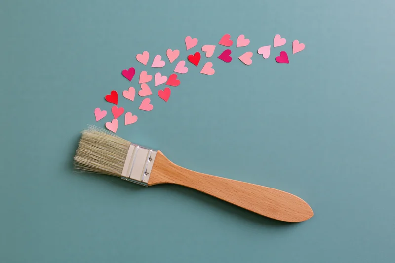 Paintbrush with wooden handle lying on teal surface, small paper hearts scattered from bristles in arc shape